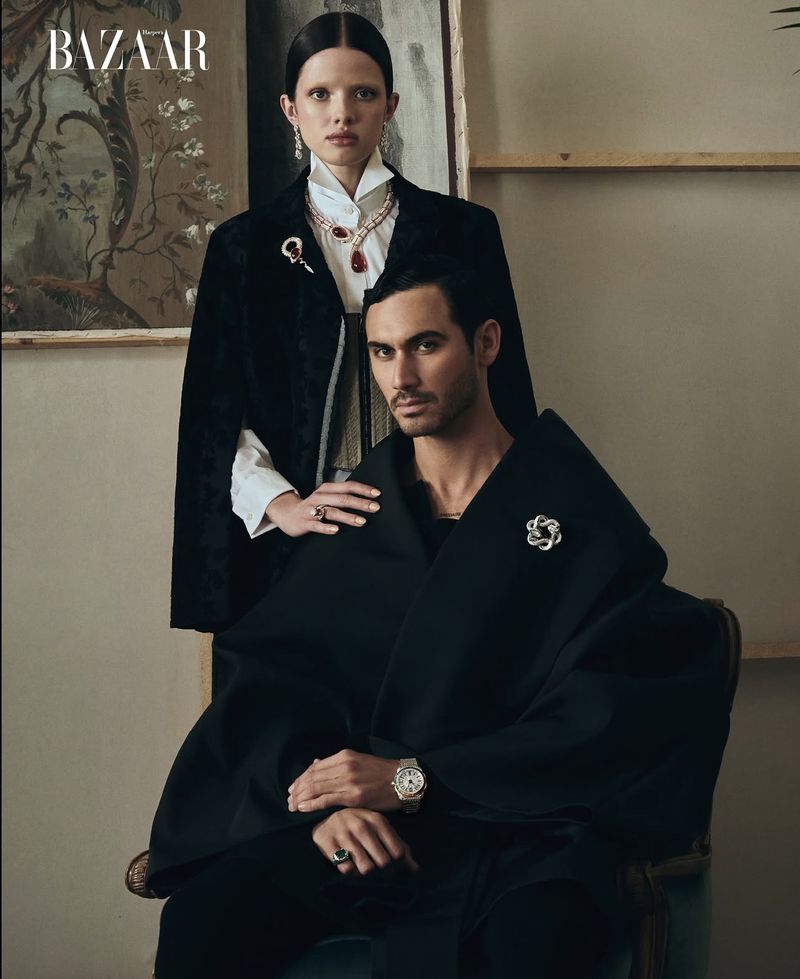 Editorial shot of a man and woman in black outfits with jewelry, posing in front of a neutral wall with a painting. Bazaar logo visible.