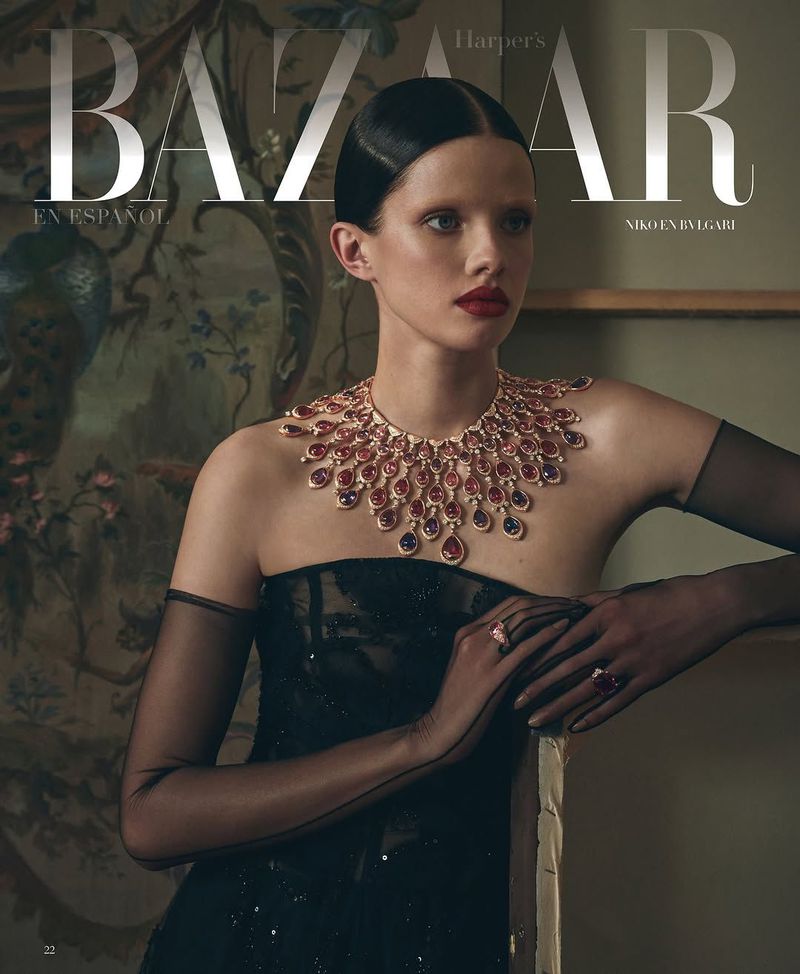 A young woman poses for a Harper's Bazaar magazine cover, wearing a black dress and statement Bulgari necklace, indoors with painting backdrop.