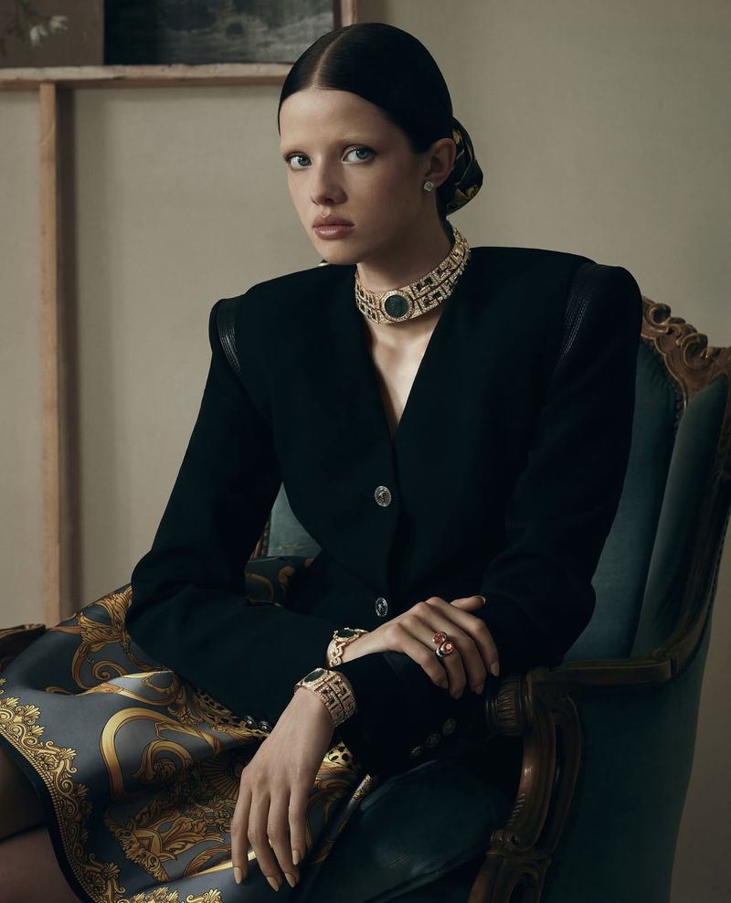 Model seated in an antique armchair wearing a tailored black jacket, statement gold choker necklace, and ornate baroque print scarf.