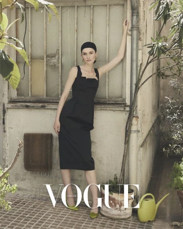 Elegant woman in a black dress, posing against a weathered wall with a window, surrounded by plants and natural light, Vogue magazine cover.