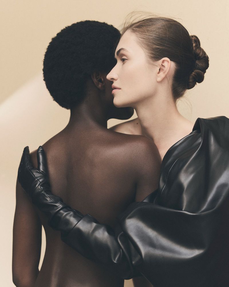 Two women in a studio setting. One with dark skin and one with light skin are in close proximity, with black gloves visible.