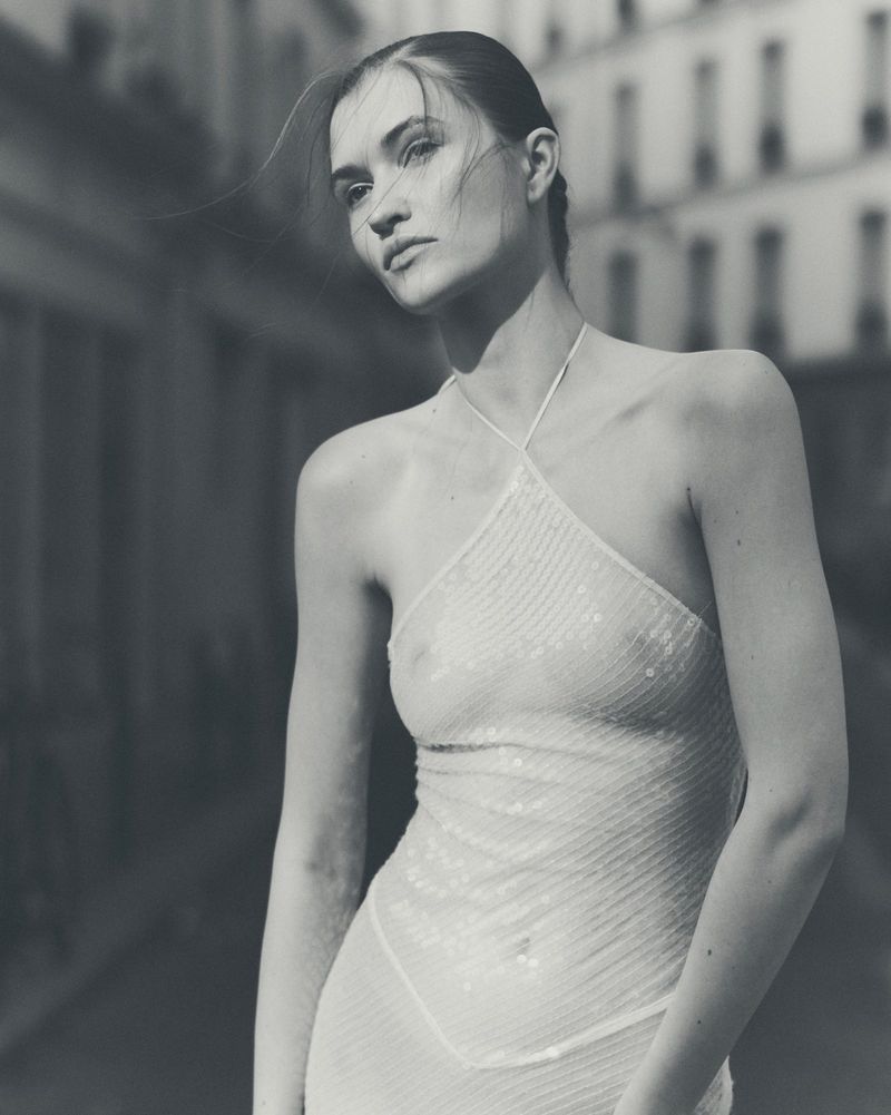 Monochrome portrait of a woman wearing a sheer halter top on a city street with soft sunlight.