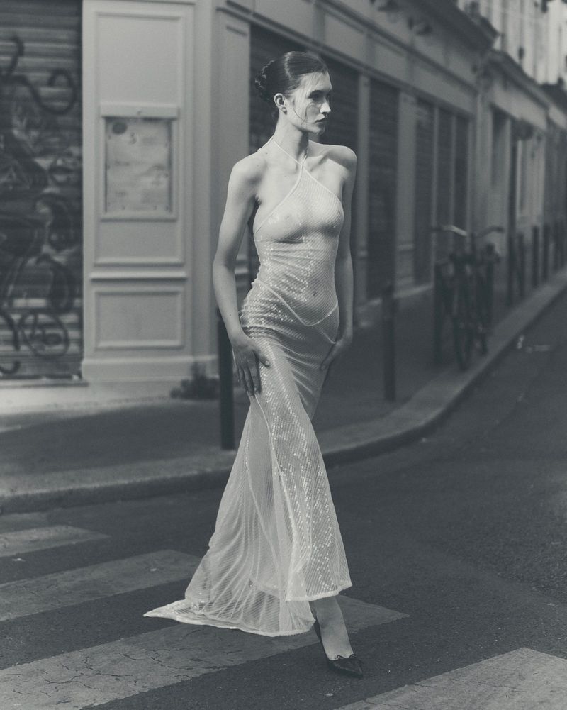 Model walking across a street crosswalk wearing a sheer sequined halterneck gown in a black and white fashion editorial.