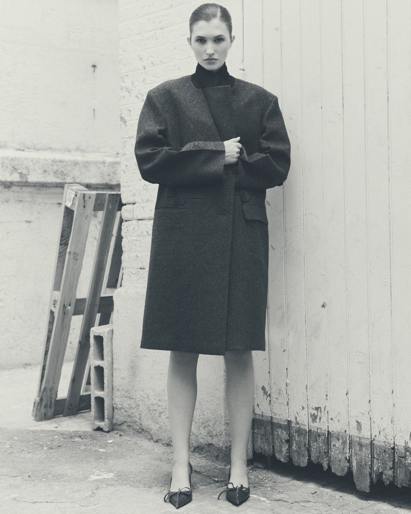 Monochrome full shot of a female model in an oversized gray coat, black top, and black heels standing against a white wall with a wood pallet.