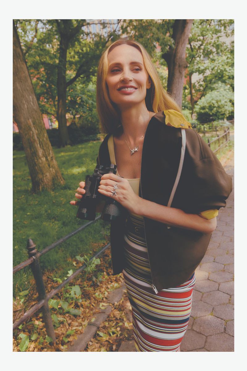 A woman smiles in a park, wearing a striped skirt and brown jacket, holding binoculars near a wrought-iron fence with leafy trees.