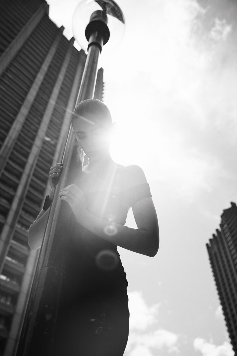 Monochrome portrait of a woman in an urban setting, standing by a street lamp with tall buildings in the background under bright sunlight.