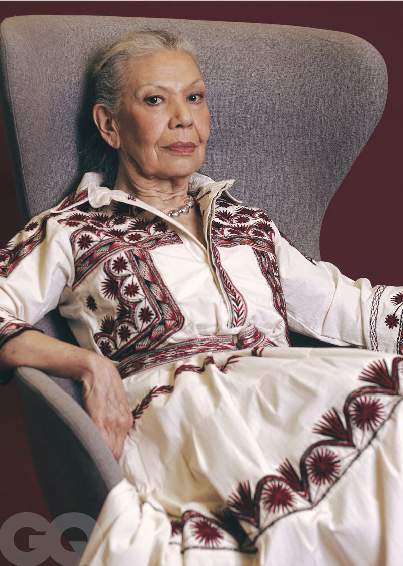Elderly woman with short gray hair in patterned white dress sitting in gray chair against a maroon background, portrait orientation.