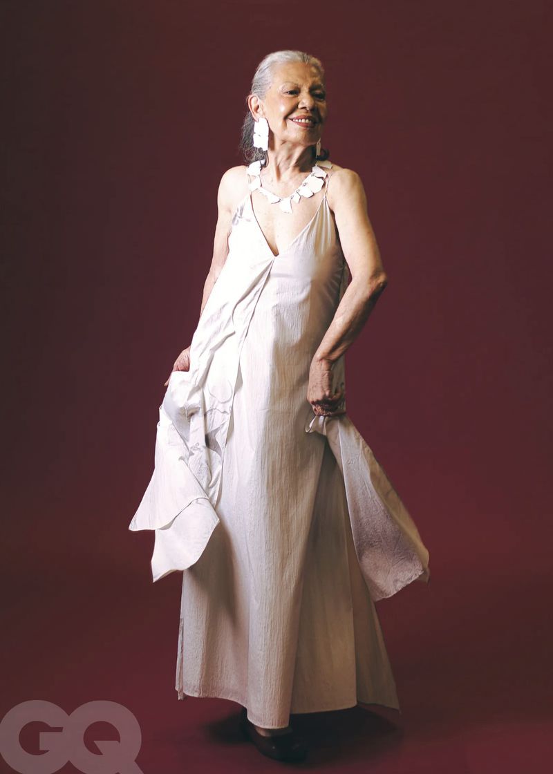 Elderly woman with white hair smiles in a full-length portrait in a white dress and statement earrings and necklace against a red background.