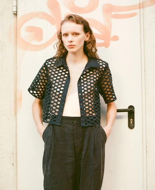 A young adult woman stands in front of a door wearing a black, short-sleeved mesh shirt, and loose black pants in a medium shot.