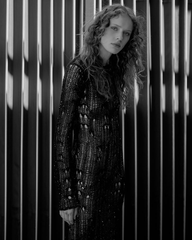 Monochrome portrait of a woman with long, wavy hair wearing a dark knit dress, standing against a striped background with vertical lines.