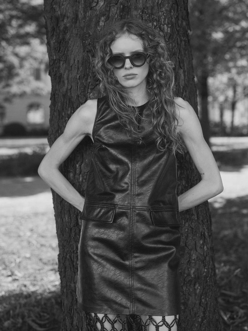 Model with wavy hair wearing dark sunglasses and a sleeveless black leather dress posing against a tree trunk in black and white.