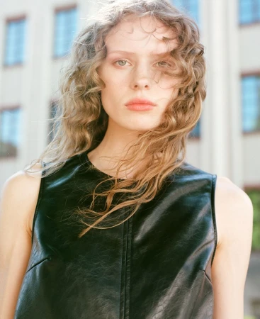 Close-up portrait of a woman with curly hair wearing a sleeveless black leather top with an out-of-focus building behind her.