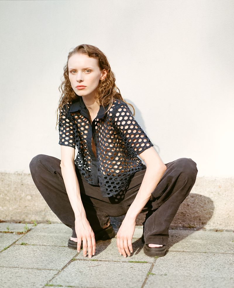 A slender female model squats on a stone surface with a white wall behind her, showcasing a sheer black, polka dot shirt and black pants.