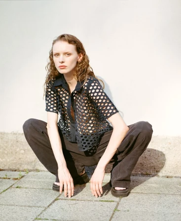 A slender female model squats on a stone surface with a white wall behind her, showcasing a sheer black, polka dot shirt and black pants.