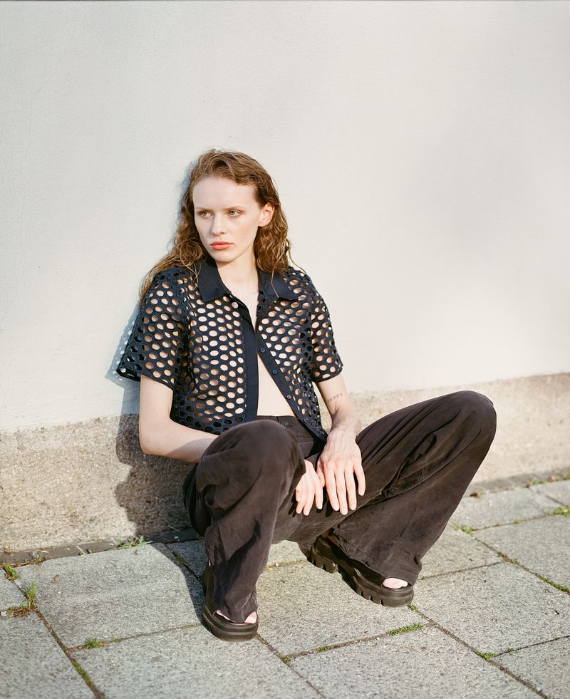 Young adult woman with wavy hair poses in a black mesh shirt and loose pants, sitting against a neutral wall and gray sidewalk.