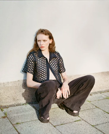 Young adult woman with wavy hair poses in a black mesh shirt and loose pants, sitting against a neutral wall and gray sidewalk.