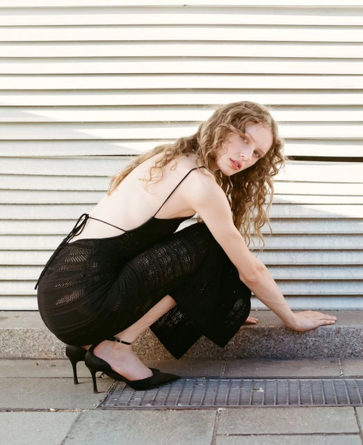 Young woman with curly hair wearing a black dress and heels is crouching on a city street, posing in front of a white wall.