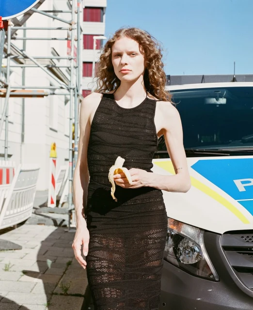 Woman in black dress holds banana, standing in front of police van on city street. Bright daylight, medium shot, urban setting.