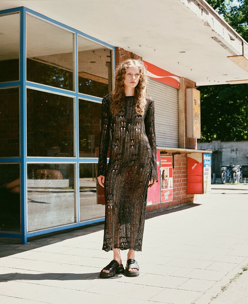 A model poses in a long-sleeved sheer maxi dress and platform sandals in front of a closed concession stand on a sunny day.
