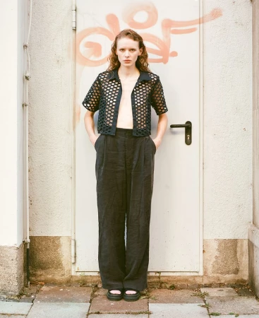 Model standing straight against a closed metal door with orange spray paint graffiti, wearing a black mesh patterned short-sleeve shirt and dark trousers.