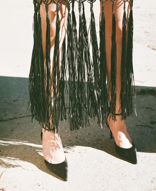 A model showcases a black tassel dress and high heels, casting shadows in an outdoor setting with diffused daylight.