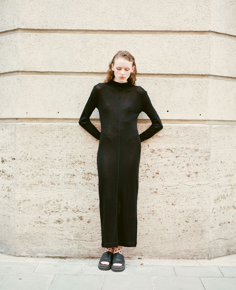 Elegant, slender woman in a long-sleeved black maxi dress stands against a textured stone wall. She has her hands on her lower back.