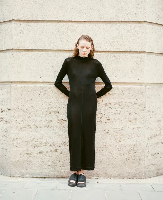 Elegant, slender woman in a long-sleeved black maxi dress stands against a textured stone wall. She has her hands on her lower back.