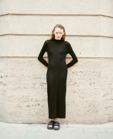 Elegant, slender woman in a long-sleeved black maxi dress stands against a textured stone wall. She has her hands on her lower back.