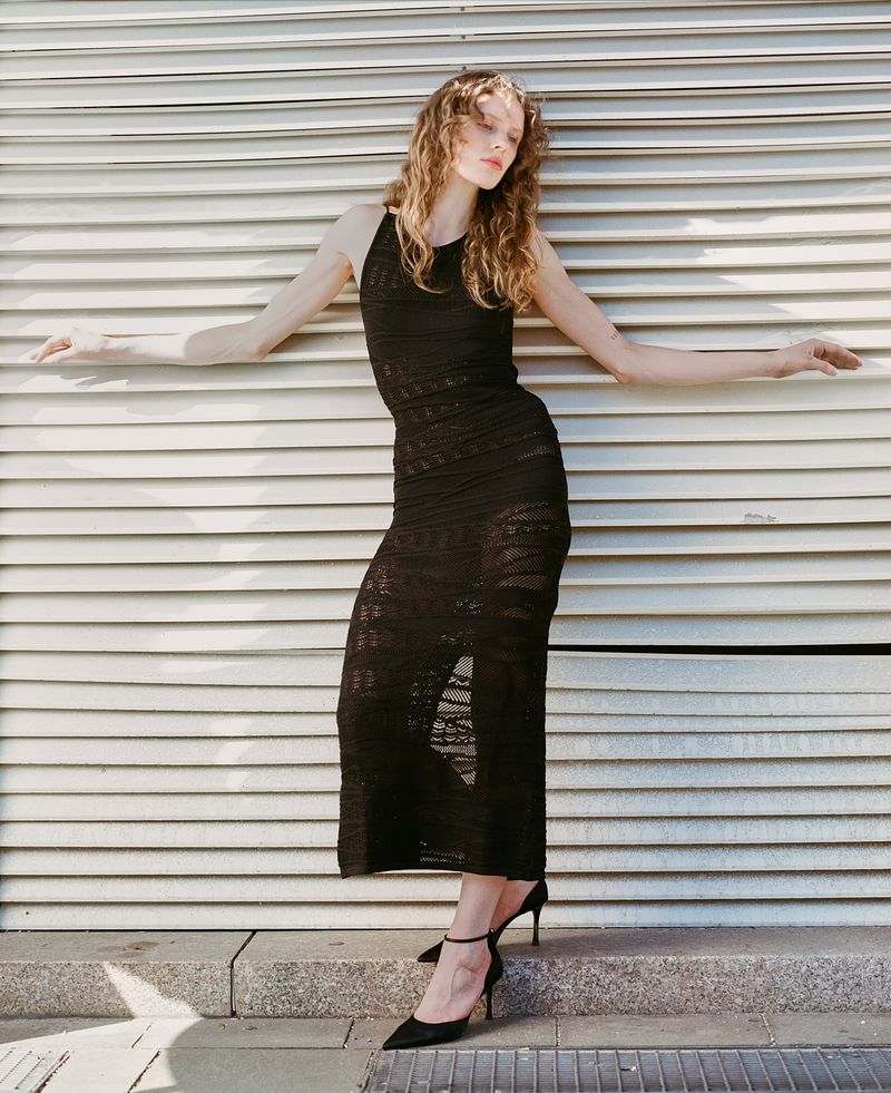 A slender woman in a black dress poses with arms outstretched against a horizontal corrugated metal wall outdoors in natural light.