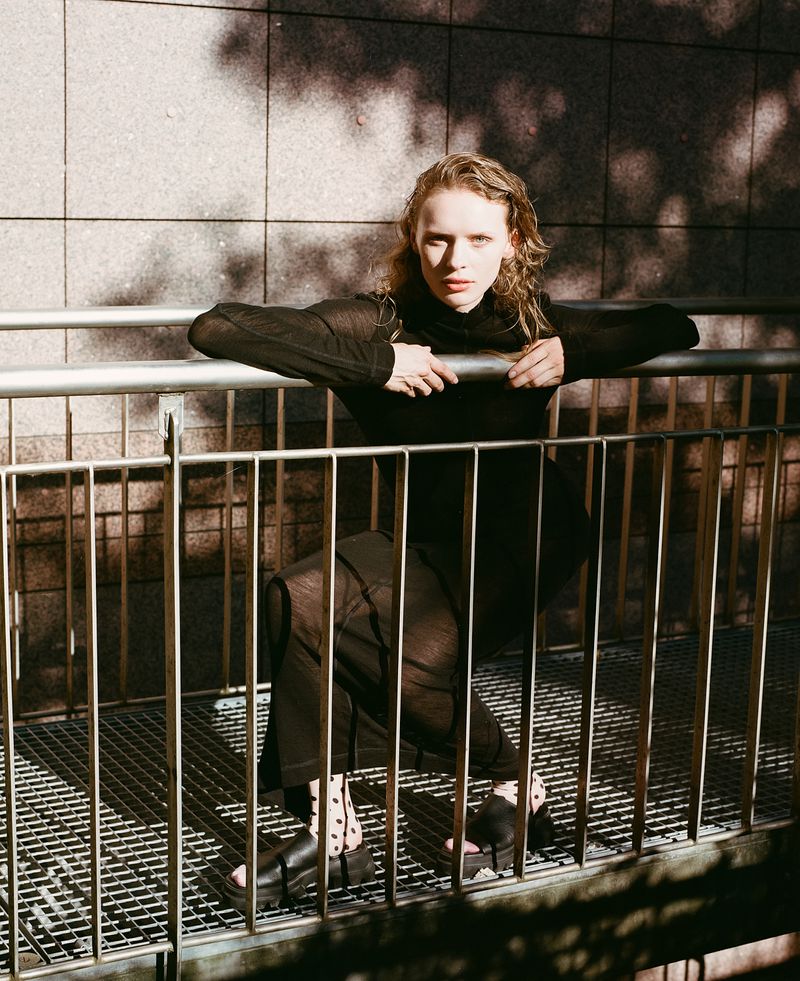 A fair-skinned young adult woman with long wavy blonde hair and black clothes poses behind a railing against a concrete background.