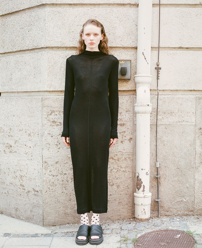 A slender woman in a black dress stands against a textured wall with a white pipe. She's wearing patterned socks and platform sandals.