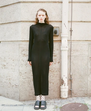 A slender woman in a black dress stands against a textured wall with a white pipe. She's wearing patterned socks and platform sandals.