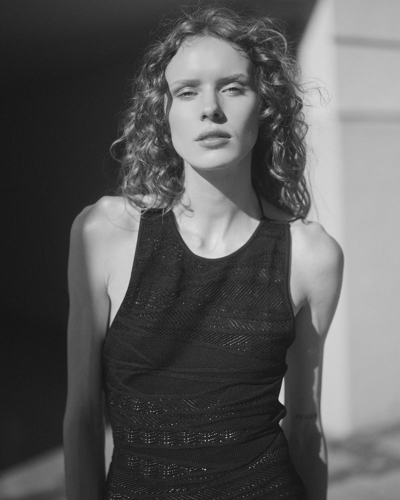Black and white portrait of a woman with curly hair wearing a textured black sleeveless top in direct sunlight.