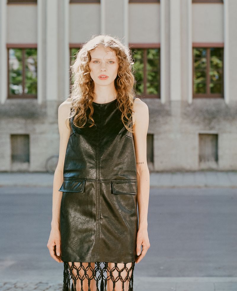 A long-haired woman in a black dress stands outside a building with large windows, in a medium eye-level shot with natural lighting.