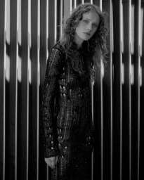 Monochrome portrait of a woman with curly hair wearing a black open-knit long-sleeve dress against a background of vertical slats.