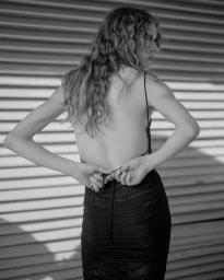 Monochrome shot of a woman with wavy hair seen from behind tying the straps of a black backless knit dress against a slatted shutter.
