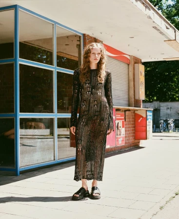 Model wearing a long black distressed knit dress and black platform slides standing outside a vintage kiosk.
