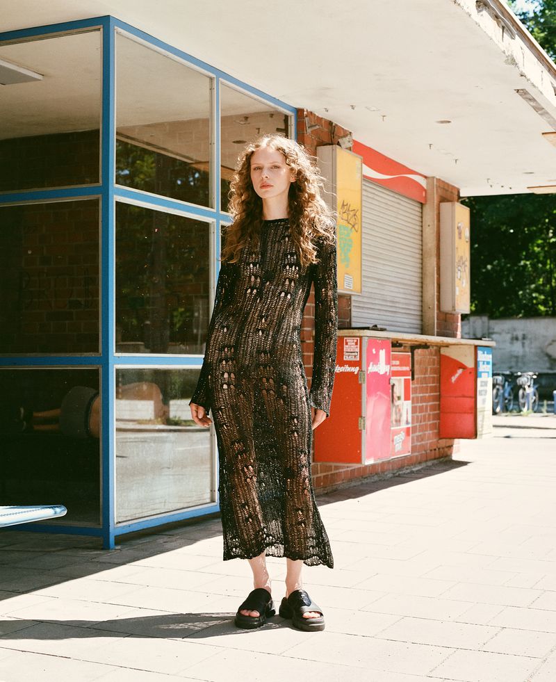 Woman with curly hair standing on a sunlit pavement wearing a sheer black open-knit maxi dress and black sandals.