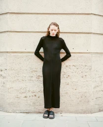 Model standing against a concrete wall wearing a long black dress and dark sandals
