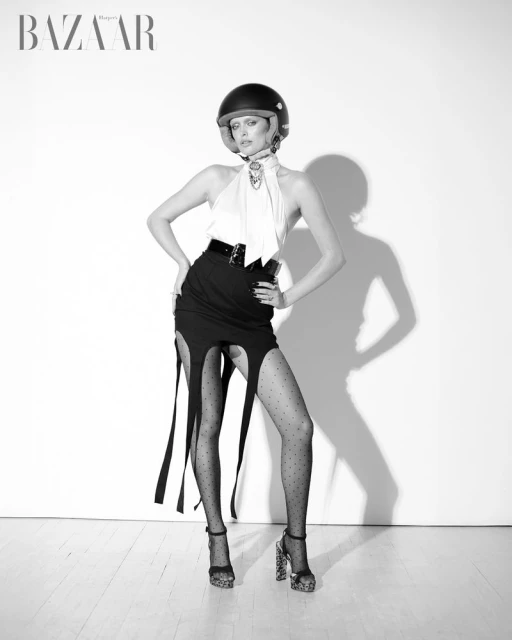 Black-and-white studio shot of a female fashion model in black skirt and polka dot hosiery, posing with a motorcycle helmet.