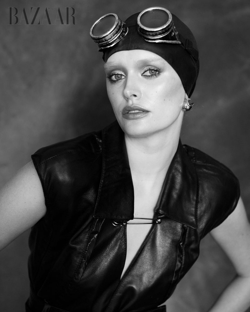 Black and white portrait of a woman wearing leather vest and cap with goggles, posing against a mottled grey background in a studio.