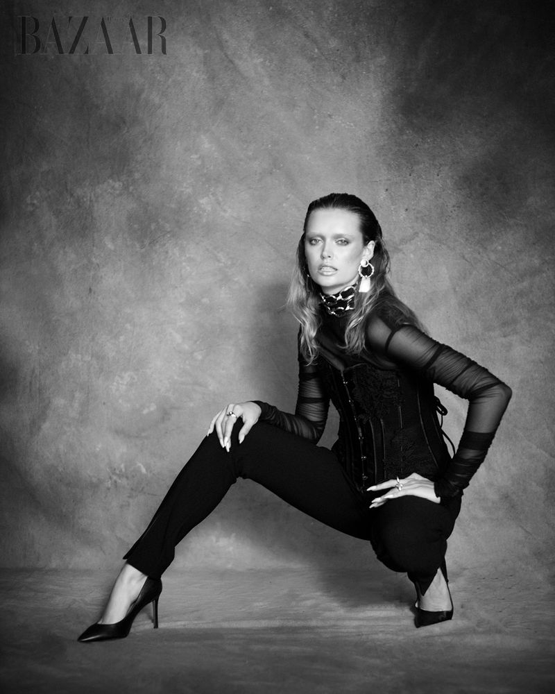 Black-and-white fashion shot of a stylish woman in a corset, pants, and high heels, crouching in a photo studio, with Bazaar logo.