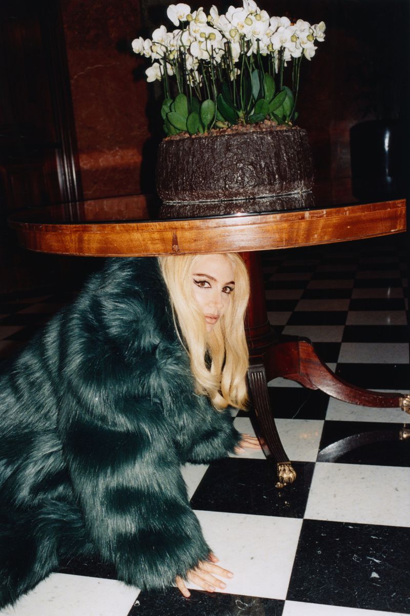 Adult female in a blue faux fur coat poses under a round wooden table topped with a potted arrangement of white orchids indoors.