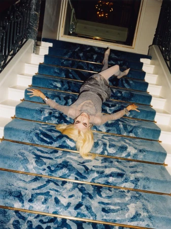 Woman lying on a blue patterned carpeted staircase. She is wearing a sparkly dress and sheer stockings and the staircase has brass rails.
