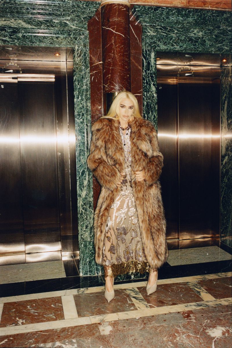 A woman with blonde hair wearing a long fur coat and dress poses indoors near two elevator doors with marble accents.