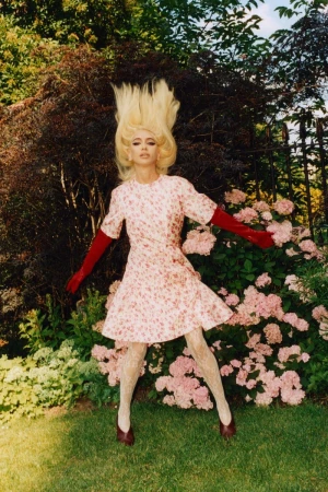 Fashion model in a floral dress, red gloves, and white patterned tights, standing in a garden with lush greenery and pink flowers.