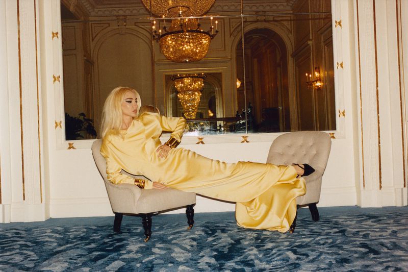 A woman with platinum hair poses in a yellow satin dress and gold cuffs while lounging on a gray armchair in a mirrored room.