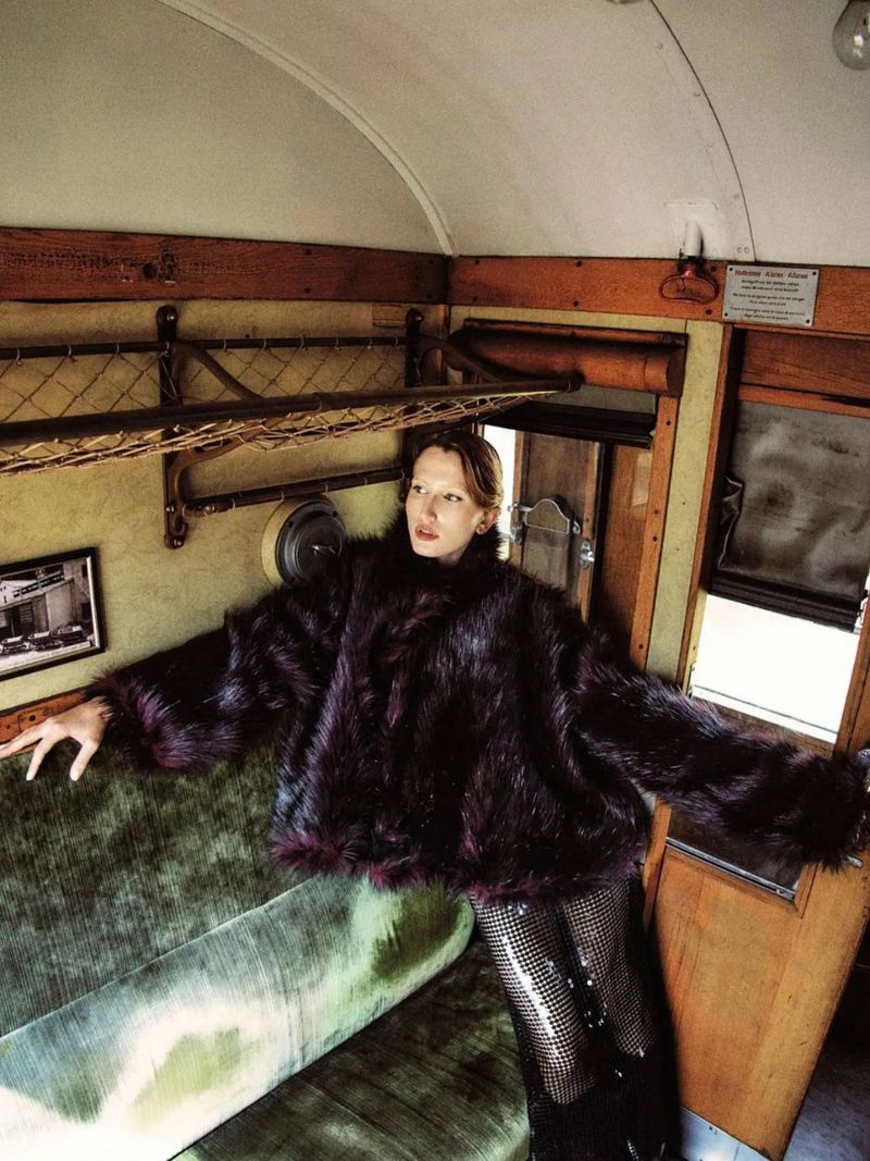 A woman in a dark fur coat poses on a green velvet seat inside a vintage train car, creating a retro fashion editorial scene.