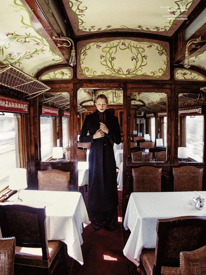 A slender woman in a long black coat stands in a vintage train dining car with white-clothed tables, wood paneling, and floral-painted ceiling.