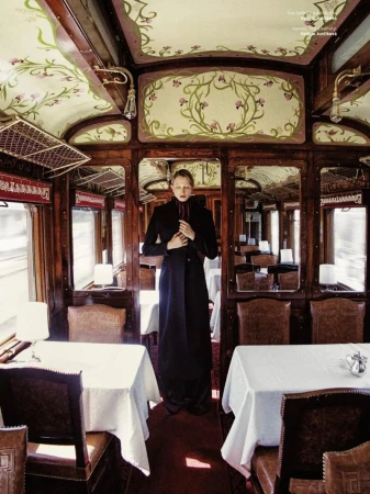 A slender woman in a long black coat stands in a vintage train dining car with white-clothed tables, wood paneling, and floral-painted ceiling.